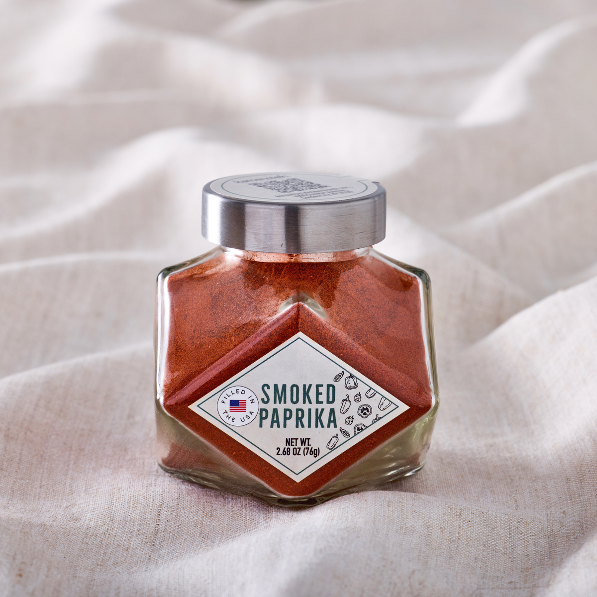 Smoked paprika in a clear diamond shaped jar with a silver lid on a soft beige fabric background