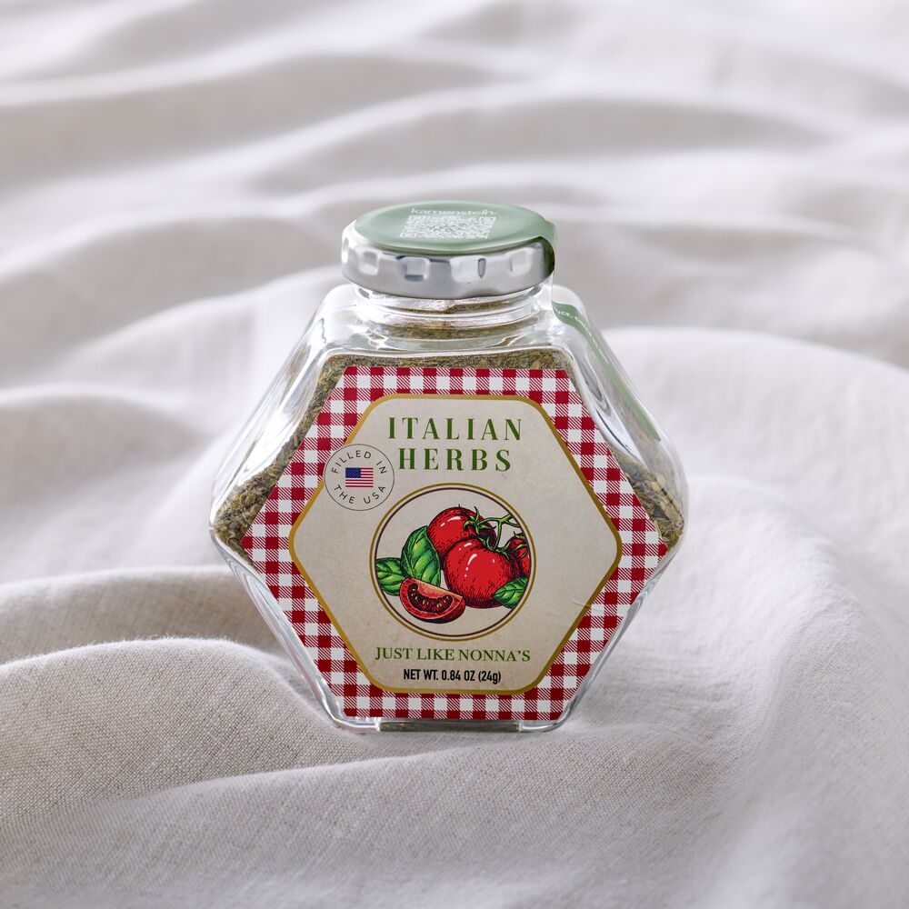 A hexagonal glass jar of Italian Herbs with a red and white gingham label and a green lid, set on a soft white fabric background.