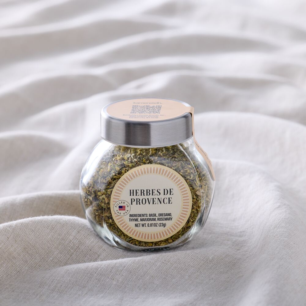 A clear glass jar filled with dried herbs, featuring a silver metal lid and a beige circular label that reads 'HERBES DE PROVENCE', set against a draped white fabric background.