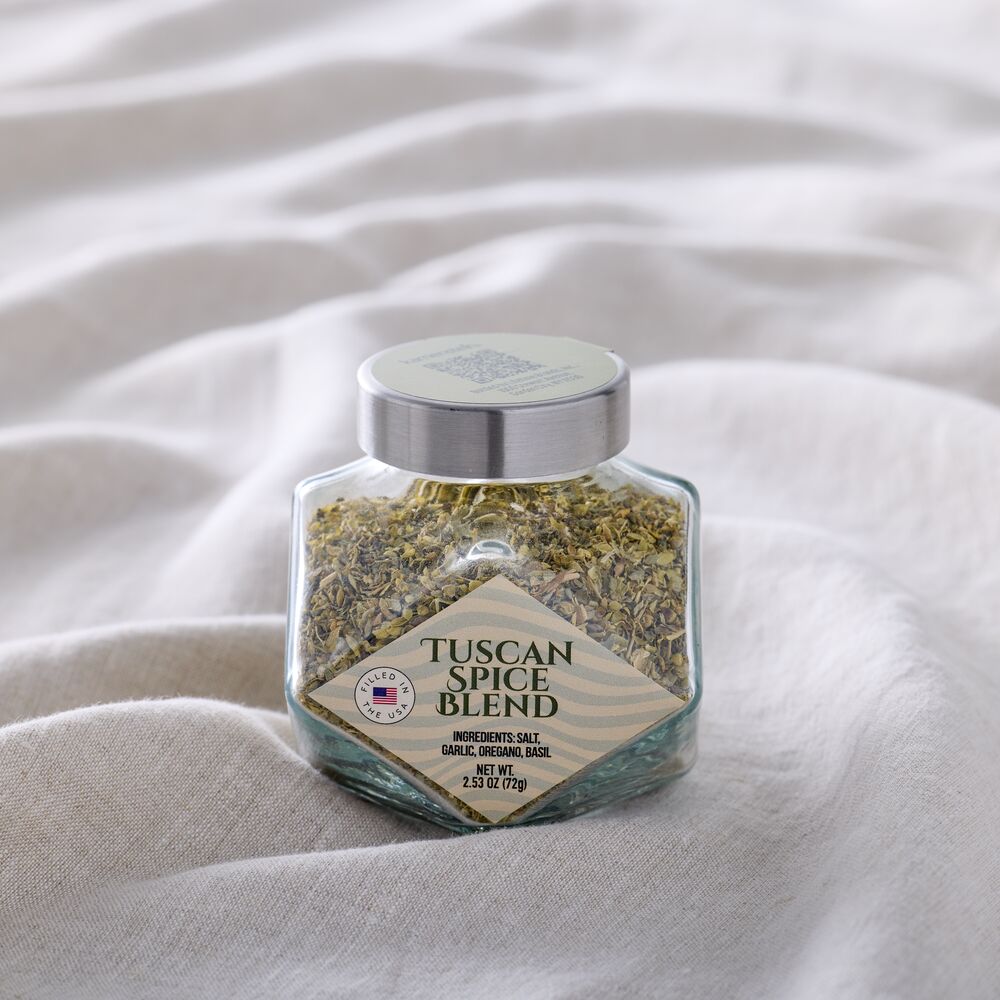 A hexagonal glass jar of Tuscan Spice Blend garlic and onion salt, filled with dried herbs and spices, resting on a draped beige linen fabric background.