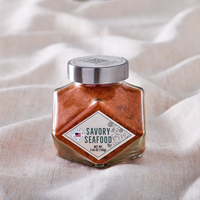 A glass jar of Savory Seafood seasoning blend with a silver lid, set against a draped beige linen fabric background.